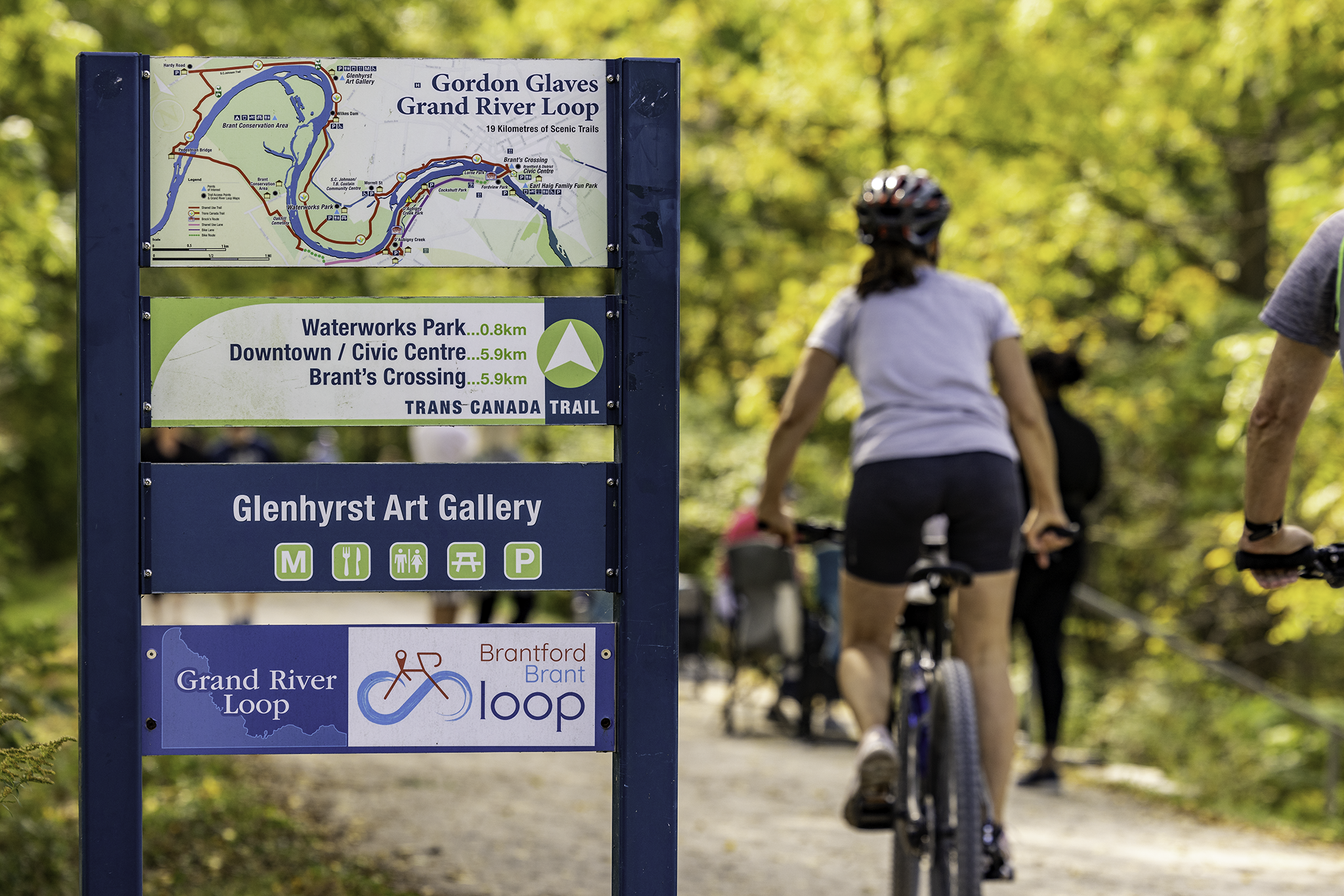 A woman rides a bike away from the camera, on a paved trail in the woods. A trail map is visible next to her, with a small sign on it that says "Grand River Loop."