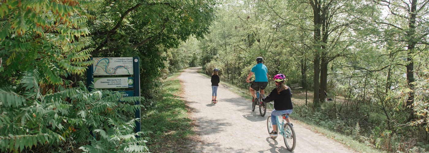 Cyclists riding on a trail in the woods - a man on a bike, a girl on a bike, and a girl ahead of them on a scooter or rollerblades. A trail sign is visible beside them.